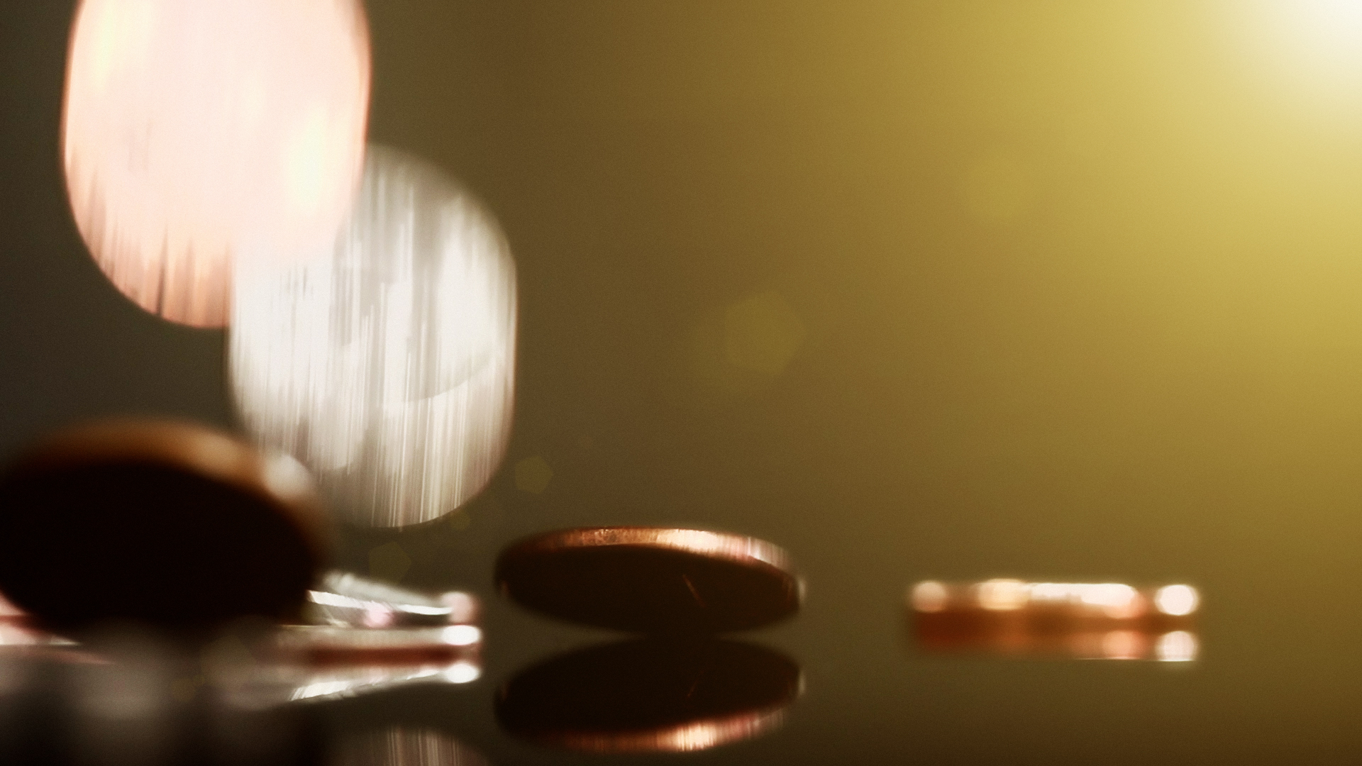 Silver and copper coins falling onto a reflective surface, showing  motion blur and lens flare. Copy space on the greeny-gold background.